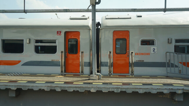 economy class train cars on station platform, sleek silver exterior accented with orange doors, metal support pole and overhead electrical wiring, modern rail transport     