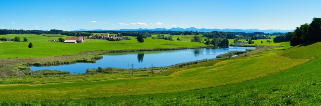Idyllic Bavarian pre-Alpine landscape with a small lake and mountains in the background on a late summer day with blue skies. 3x1 panoramic photo.
