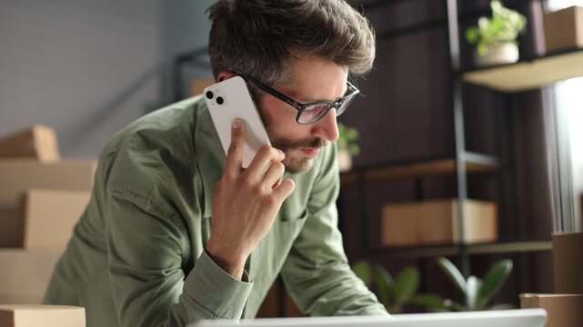 A man is talking on his cell phone while sitting at a desk with a stack of cardb