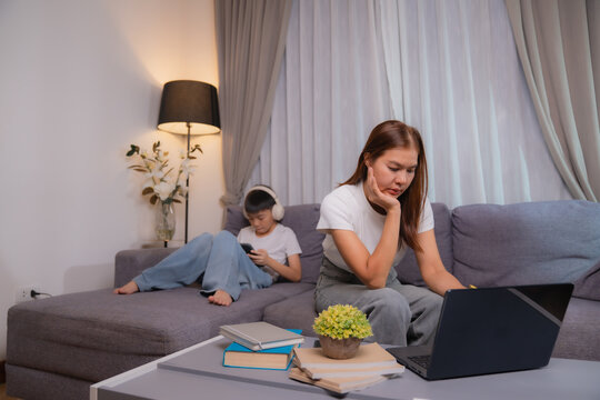 Asian businesswoman is working using laptop and feeling stressed while her son is lying on the couch and playing video games on smartphone at home