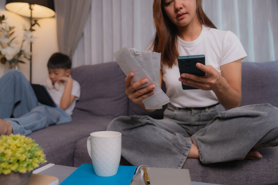Young stressed asian mother calculating domestic bills using smartphone and calculator sitting on the sofa in living room at home with her son playing with tablet in the background