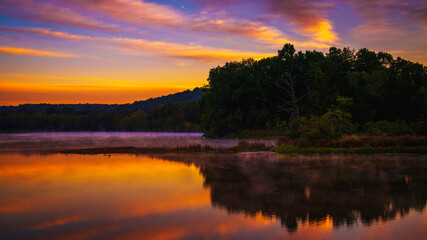 Sunrise lakeshore autumn landscape with forest, colorful clouds, and dawn sky reflected on pond water, a tranquil glimpse of Connecticut’s natural beauty in the heart of New England.