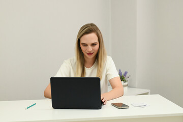 The photo shows a young woman sitting at a table with a laptop and smartphone in her hands.