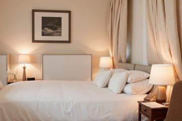 Neatly made bed with bedding standing in softly lit bedroom, bedside tables holding lamps and telephone, framed black and white photograph hanging above headboard