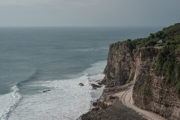 A stunning view of the rugged cliffs and coastline of Uluwatu, Bali. A winding pathway follows the edge of the powerful Indian Ocean waves below.

