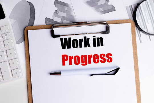 Clipboard with paper showing the words work in progress, along with a pen, calculator, charts, 