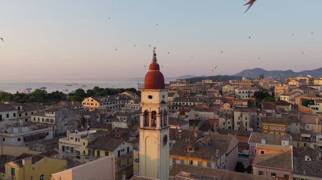 Saint Spyridon Church and bell tower in Old Town Corfu under warm morning sunrise, swallows swirl in sky above, aerial drone orbit