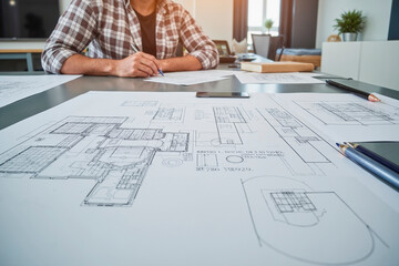 Caucasian young adult man working on architectural blueprints at desk, holding pen and reviewing technical drawings, smartphone and drafting tools visible on workspace