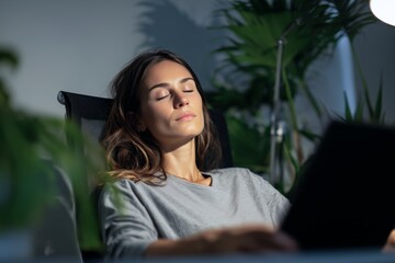 Exhausted female entrepreneur closing her eyes in office chair, moment of stillness during late night work session, surrounded by green plants