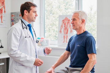 Caucasian young adult male doctor consulting middle aged Caucasian man sitting on examination table in medical office, both engaged in conversation during health checkup
