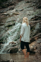 Young woman with long hair stands with her back to the camera, gazing at a mountain waterfall. The...