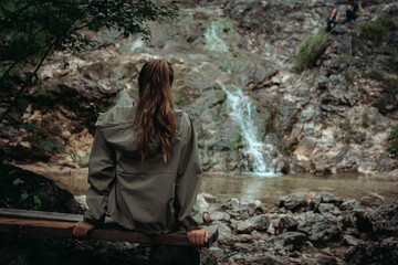 Young woman with long hair stands with her back to the camera, gazing at a mountain waterfall. The...