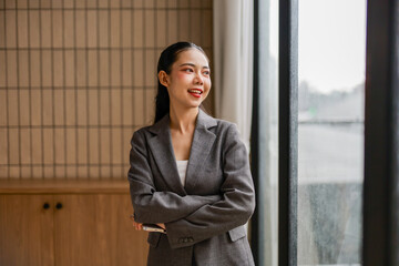 Young Asian businesswoman holding a smartphone, gazing out of the office window, savoring her...