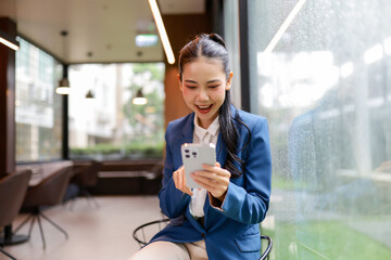 Asian businesswoman expressing excitement while using smartphone in modern office, enjoying mobile...