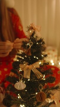 Vertical video. Woman in Red Pajamas Placing Ornament on Top of Mini Christmas Tree with Golden Bows and Lights in Cozy Night Setting