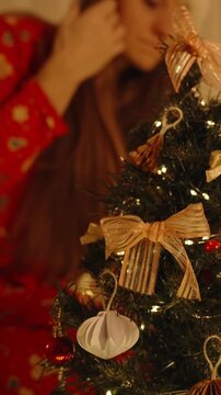 Vertical video. Woman in Santa Hat Bending Toward Mini Tree and Holding Ornament in Hand Surrounded by Warm Lights and Presents