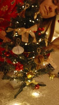 Vertical video. Close Up of Woman&rsquo;s Hand Adjusting Gold Ribbon and Ornaments on Mini Tree with Warm White Lights in Cozy Room