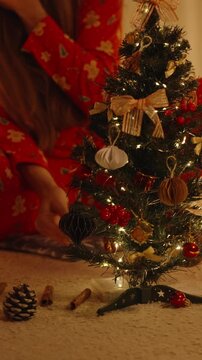 Vertical video. Woman in Red Pajamas Sitting with Decorated Mini Christmas Tree Surrounded by Gifts and Pinecones on Cozy Carpet
