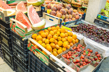 Colorful fresh fruit display at local market featuring sliced watermelon, oranges, strawberries, peaches and cherries arranged in wooden crates and plastic containers.