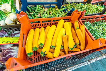 Bright yellow zucchini fill an orange crate, positioned next to other green vegetables like zucchini and long beans, along with radicchio and price tags at a bustling market.