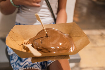 A close-up shot of a child holding a cardboard tray with a stack of pancakes drenched in smooth, melted chocolate and served with a small wooden fork.