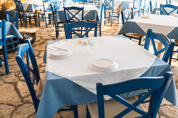 Authentic Greek taverna terrace with classic blue and white table settings, stone flooring, and traditional wooden chairs awaiting seaside diners.