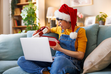Woman wearing Santa hat unboxing Christmas present while having video call using laptop computer
