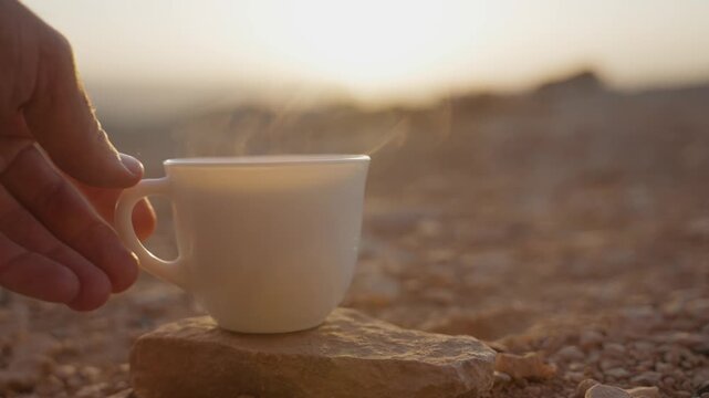 Hand setting hot drink cup with steaming tea or coffee on desert rock or stone. Early morning sunrise or sunset coffee break during road trip in Middle East. Enjoying stunning views outdoors in nature