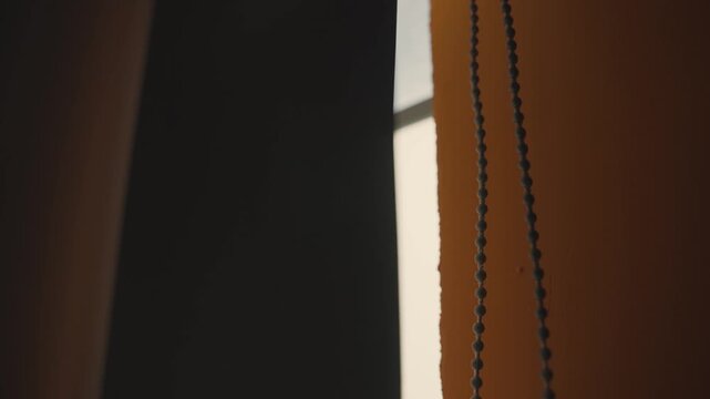 Extreme close up of a roller blind slowly moving in the wind, with warm sunlight peeking through the side, creating an intimate and cozy atmosphere in a modern apartment interior
