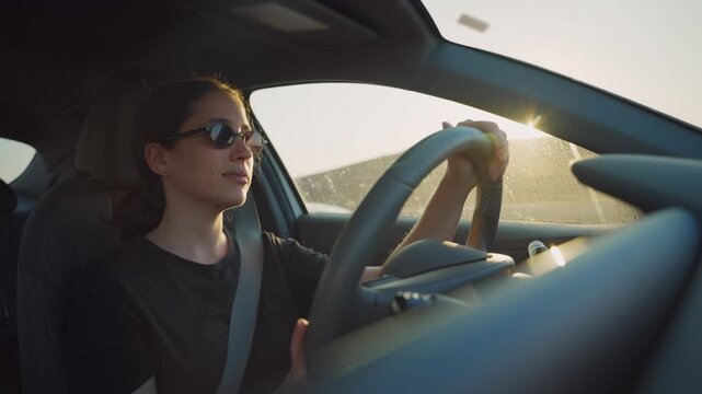 Young woman with sunglasses driving car on highway during sunset or sunrise. Female commuter going to work or leaving for vacation. Early morning sunlight or afternoon orange color. Enjoying smiling