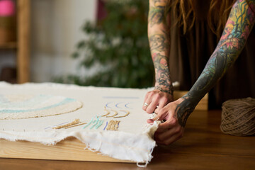Young adult Caucasian woman with tattooed arms working on handmade textile piece in carpet workshop, carefully stretching fabric over wooden frame, spool of twine nearby