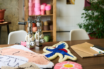 Tufting machine standing on wooden table surrounded by colorful handmade carpet samples including bird and flower motifs, sketchbook and markers visible in carpet workshop setting