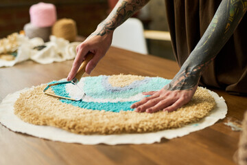 Tattooed young adult Caucasian woman using rug carving tool shaping colorful handmade carpet on wooden table in carpet workshop, focusing on detailed craftsmanship with hands visible