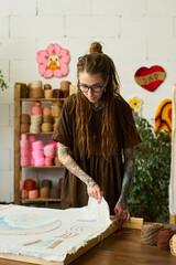 Young adult Caucasian woman with long dreadlocks and tattoos working on handmade carpet in...
