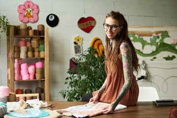 Portrait of young adult Caucasian woman with long hair and tattoos smiling at camera while working on handmade carpet in workshop, surrounded by yarn and textile materials