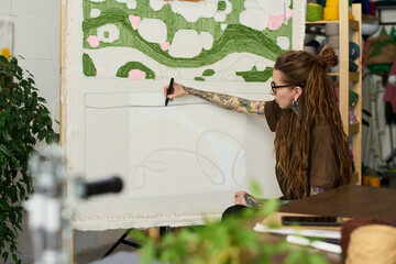 Caucasian young adult woman with long hair and tattoos working on large carpet design, sketching...