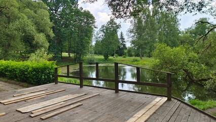 A river with algae flows among grassy meadows with growing birches, willows and other trees that are reflected in the water. A wooden pedestrian bridge with a fence spans the river. Cloudy summer