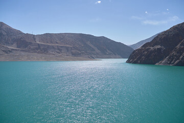 Obraz premium Sparkling Turquoise Water Reflecting Sunlight Amidst Rugged Mountain Landscape Under a Clear Blue Sky