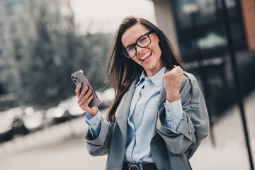 Confident young businesswoman wearing a stylish trench coat smiling outdoors while celebrating a...