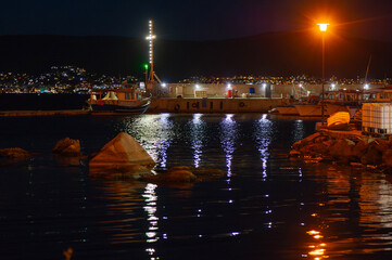 A serene harbor at night glows with reflections from docked boats and bright pier lights beneath...