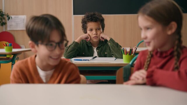 Two Caucasian children girl with boy laughing talking bullying talk about lonely sad upset African American schoolboy sitting alone racial discrimination three multiracial classmates in school lesson