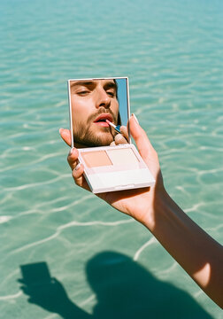 Man applying lip gloss reflected in a compact mirror held by a hand above water.
