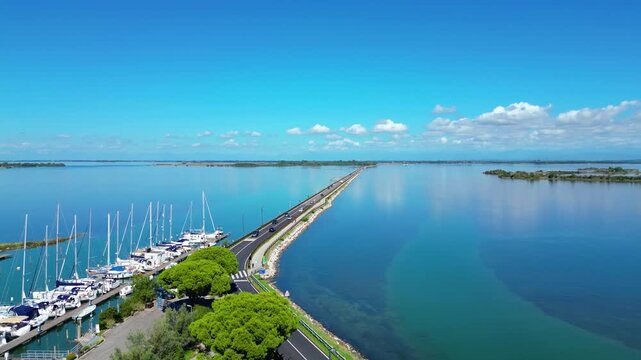 Aerial view of kayaks and boats entering Grado, Italy, with turquoise Adriatic waters, a scenic bridge, and the historic seaside town in the background. Popular destination for water sports, summer 