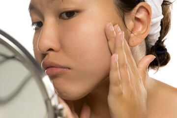 Asian woman touching her cheek while examining skin in mirror, studio shot on white background.