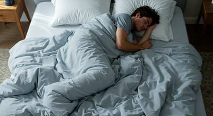 Young man sleeping peacefully in bed covered with light blue bedding during daytime rest. Sleep health concept for relaxation and bedroom comfort products