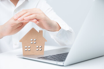 Businessman using both hands to protect a house model with a laptop beside it. Concept of property protection, real estate security, insurance, investment, and safe business planning