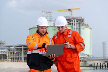 Workers collaborate on a laptop at an industrial site with storage tanks during a cloudy day