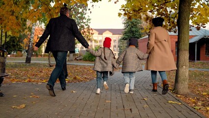 Fototapeta premium Happy family, children play, run, holding hands of mom, dad, in autumn park. Kids, mom, dad play together, holding hands in autumn outdoors. Family weekend walk, mother, child, father in park. People