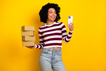Young woman holding stacked boxes with a phone in hand against a yellow background creating...