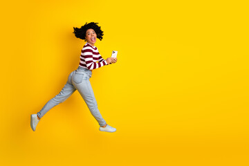 Funny woman in casual striped outfit jumping joyfully with smartphone against a vibrant yellow...